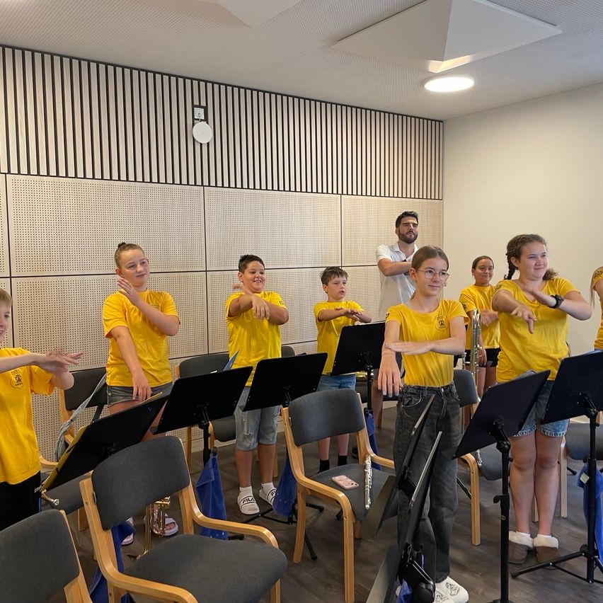 Eine Gruppe von Kindern in gelben Shirts übt Musik in einem Raum mit einem Mann, der hinter ihnen steht. Sie halten Musikinstrumente und einige lächeln.