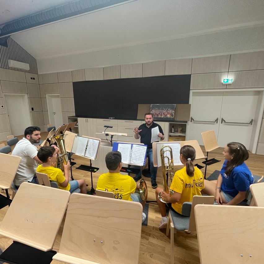 Eine Gruppe junger Musiker, die gelbe T-Shirts tragen, sitzen auf Stühlen und spielen ihre Instrumente, während ein Mann vor ihnen steht und ein Blatt Papier in der Hand hält.