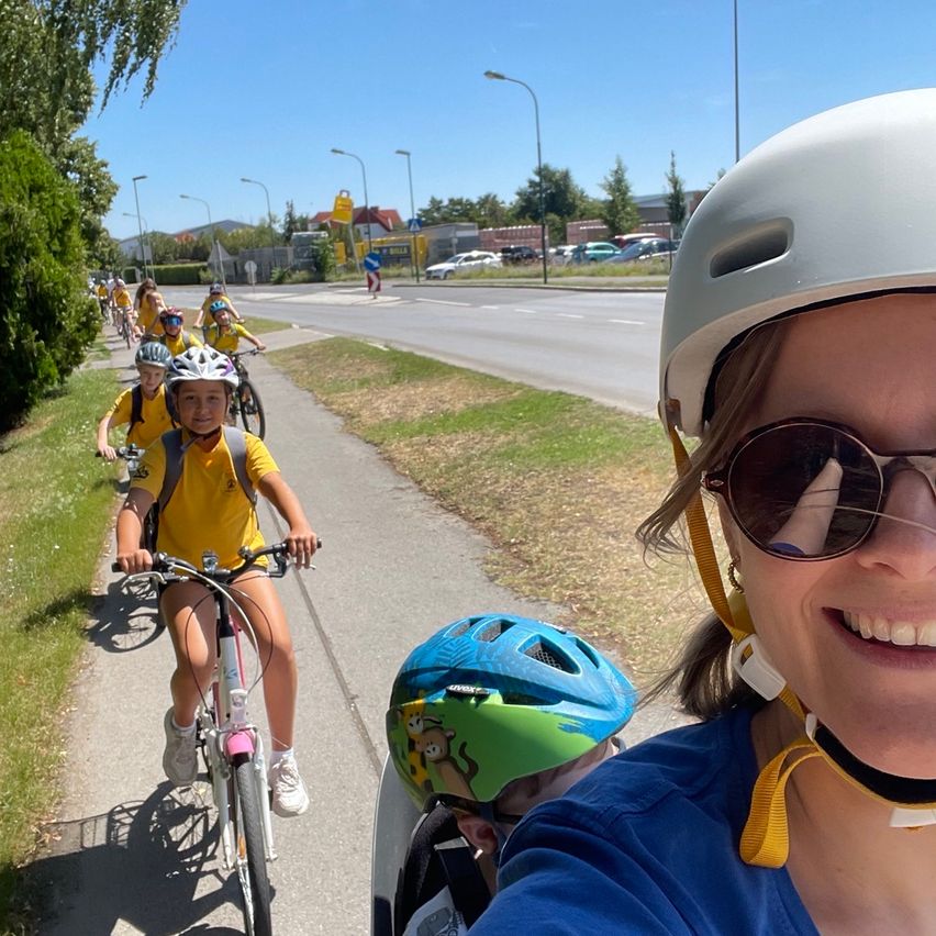 Eine Gruppe von Kindern in gelben Uniformen fährt auf einem Bürgersteig mit Fahrrädern, mit einer Frau in einem blauen Shirt und weißer Helm in der Vorderansicht.