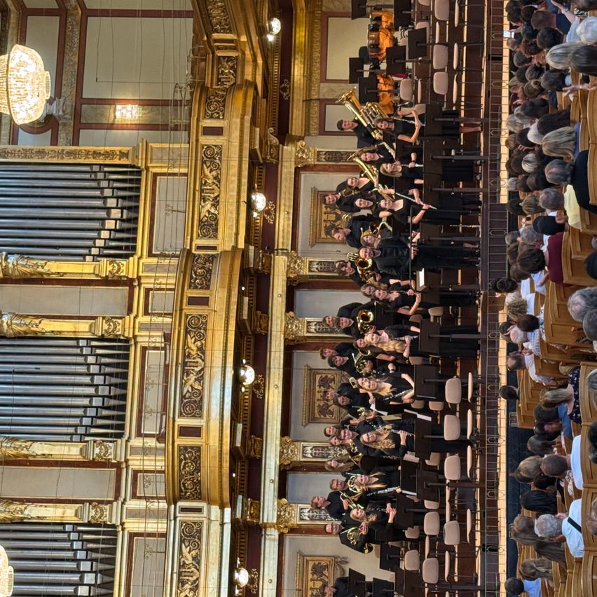 Ein großes Orchester spielt auf der Bühne in einem großen Saal, mit einer großen Orgel im Hintergrund. Das Publikum sitzt aufmerksam im Vordergrund.