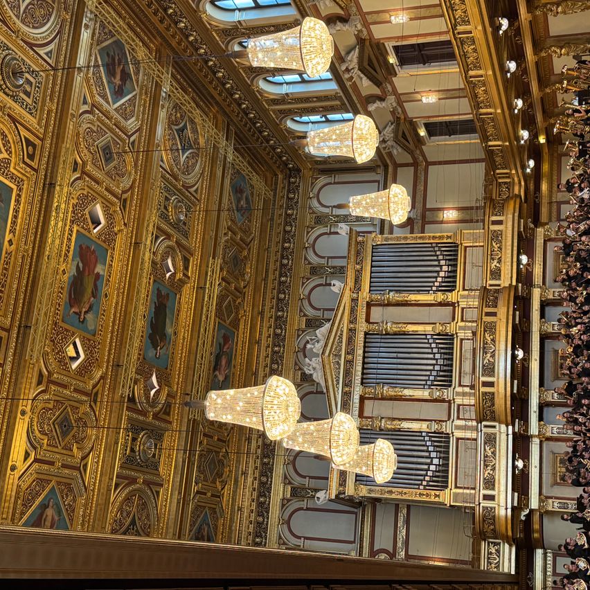 Ein großartiger Innenraum eines historischen Gebäudes mit kunstvollen goldenen Wänden, komplizierten Deckendesigns und großen Kronleuchtern, die von der Decke hängen. Eine große Orgel ist in der Mitte sichtbar, und eine Menschenmenge ist unten versammelt.