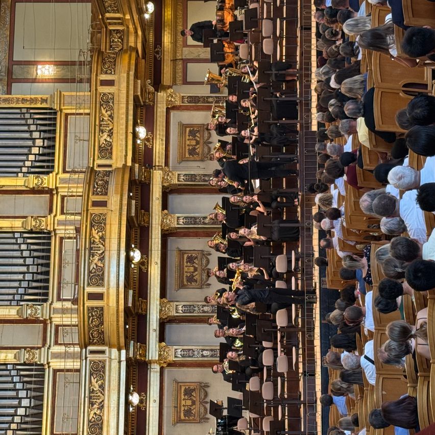Ein großes Orchester spielt in einem großen Saal, wobei die Musiker Blech- und Holzblasinstrumente spielen. Das Publikum sitzt in Reihen und beobachtet die Aufführung. Der Saal ist mit kunstvollen goldenen und weißen Dekorationen geschmückt.