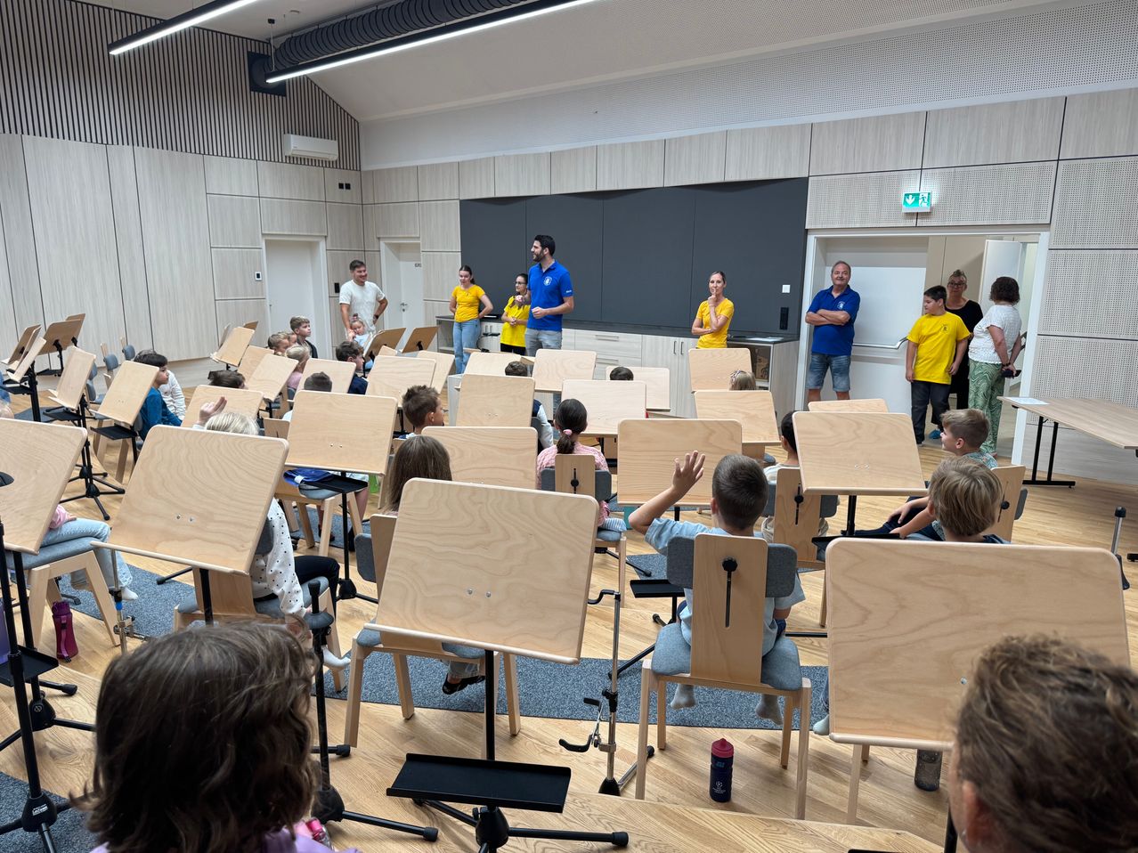 Eine Gruppe von Kindern in gelben T-Shirts sitzt in Stühlen und schaut auf eine Gruppe von Erwachsenen in einem Klassenzimmer. Die Erwachsenen stehen vor einer Tafel.