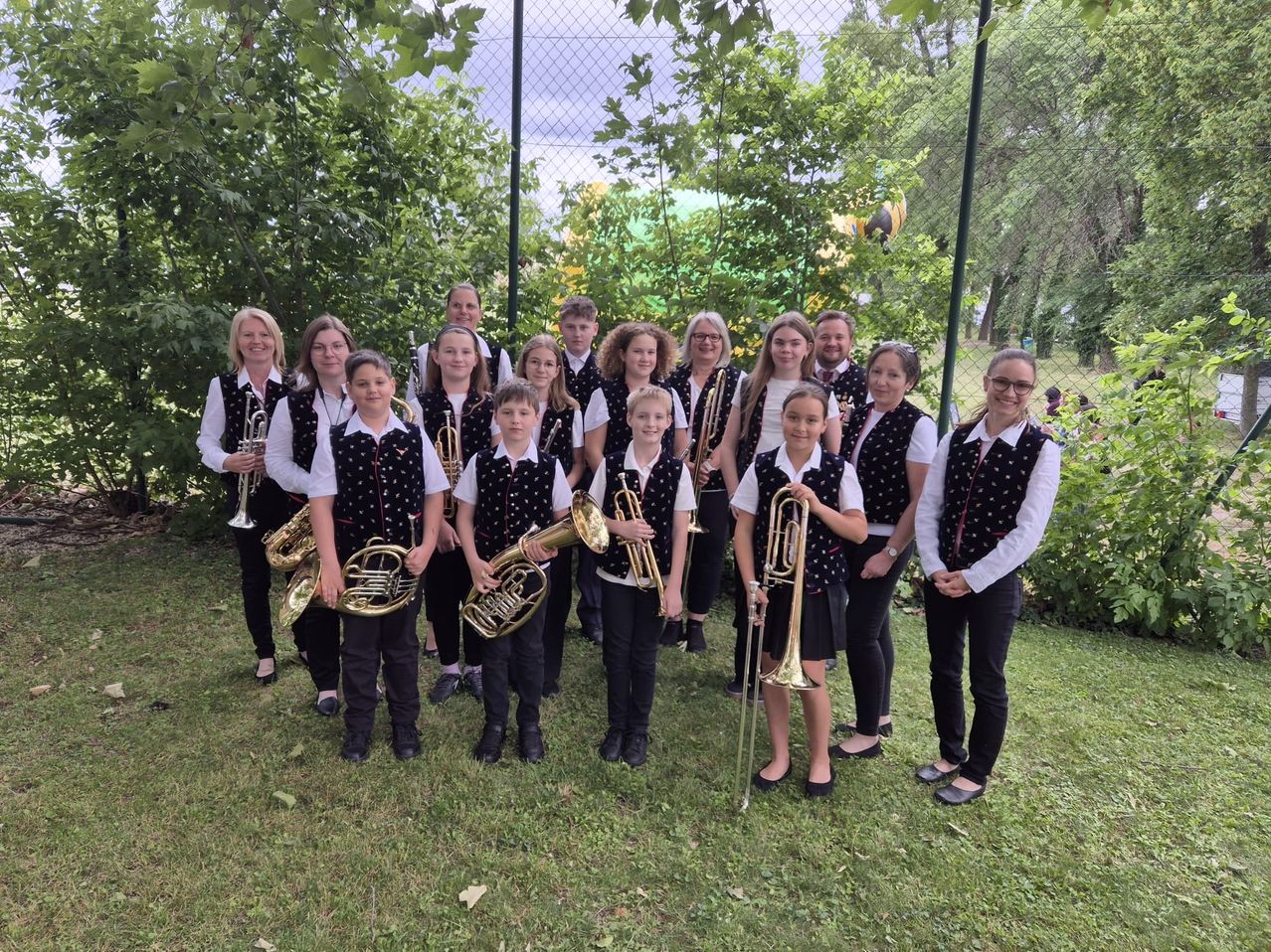 Eine Gruppe junger Musiker in einheitlichen Kostümen posiert mit ihren Blechblasinstrumenten in einem Garten mit Bäumen.