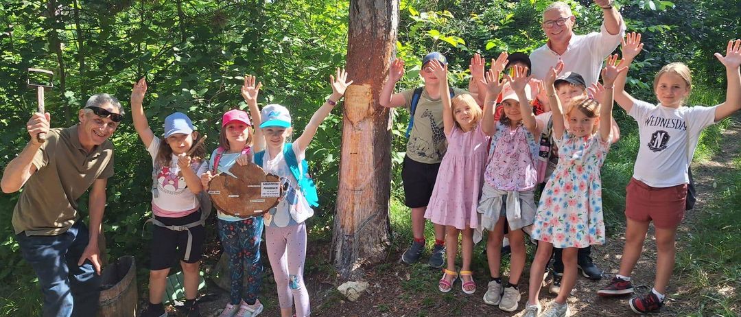 Eine Gruppe von Kindern und Erwachsenen steht um einen Baum in einem Wald, einige winken und lächeln. Ein Mann im Hintergrund gibt einen Daumen hoch.
