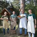 Vier Männer in Schürzen stehen in einem Wald. Einer hält einen Holzfass, ein anderer einen Teller mit Essen. Sie sind in der Nähe eines Baumstamms und eines kleinen Holzhauses.