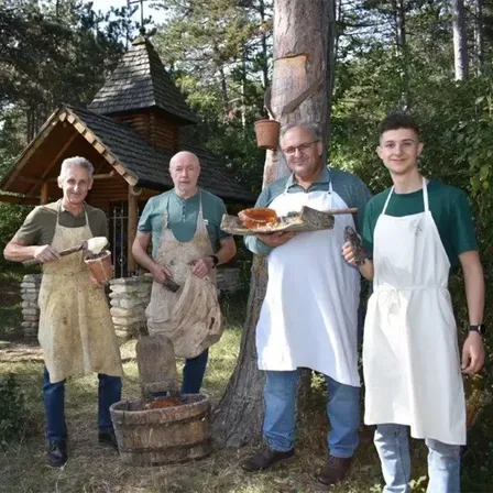 Vier Männer in Schürzen stehen in einem Wald. Einer hält einen Holzfass, ein anderer einen Teller mit Essen. Sie sind in der Nähe eines Baumstamms und eines kleinen Holzhauses.