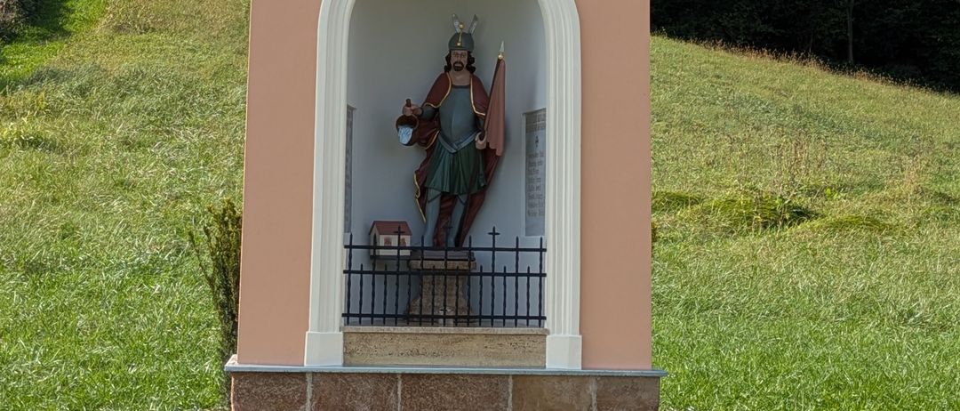 Eine kleine Kapelle mit einer Statue eines Ritters, der eine Laterne hält, befindet sich auf einem grasbewachsenen Hügel. Die Kapelle hat ein farbenfrohes Fenster und ein schwarzes Tor.