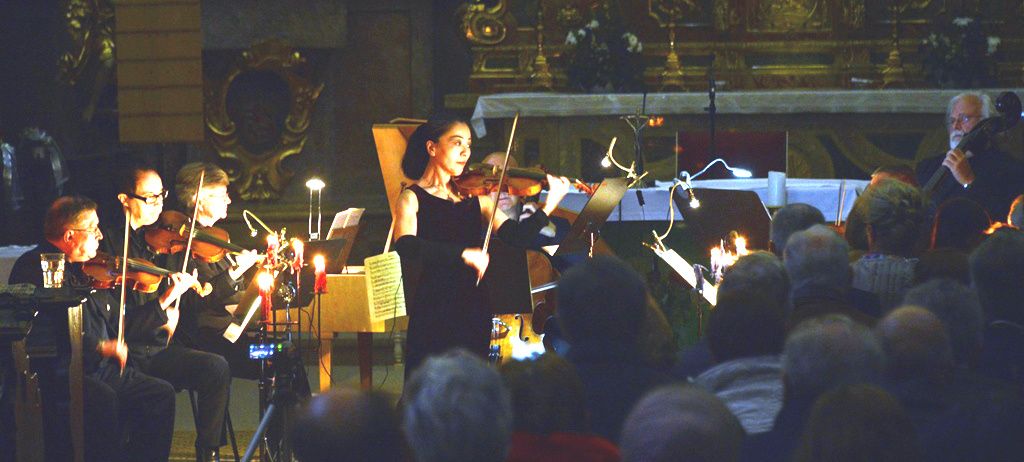 Eine Geigerin steht vor einer Gruppe von Musikern in einem schwach beleuchteten Raum. Die Geigerin ist von brennenden Kerzen und Notenständern umgeben. Das Publikum sitzt vor ihnen.