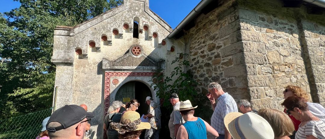 Eine Gruppe von Menschen steht vor einem alten Steingebäude mit einer Holztür und dekorativen Fenstern. Sie tragen Sommerkleidung und Hüte und schauen in Richtung des Eingangs. Bäume und ein klarer blauer Himmel sind im Hintergrund zu sehen.