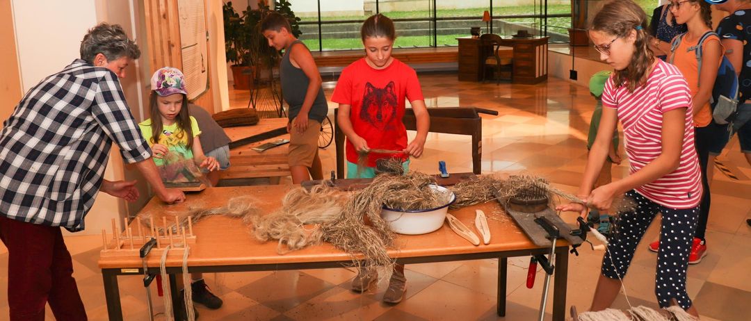 In einem geräumigen Raum mit großen Fenstern arbeiten mehrere Kinder an einem Tisch mit Stroh und Werkzeugen. Ein Mädchen in einem roten Shirt hält ein Werkzeug. In der Nähe stehen Holztische, ein Stuhl und eine Topfpflanze.