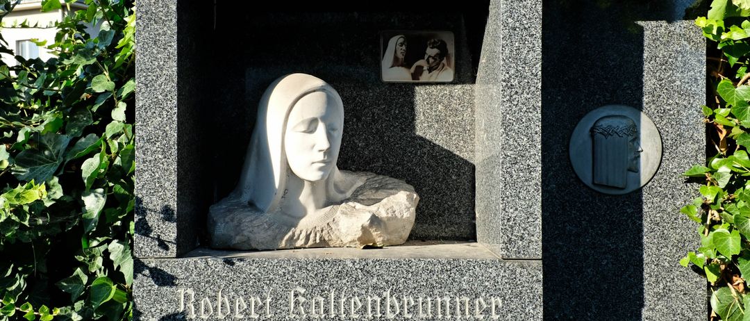 Ein Steindenkmal mit einer Statue einer Frau, den Kopf geneigt. Darunter befindet sich ein Namensschild mit der Aufschrift 'Robert Kollerbrenner 1910-1938'. Über der Statue befindet sich ein kleines Foto eines Mannes und einer Frau.