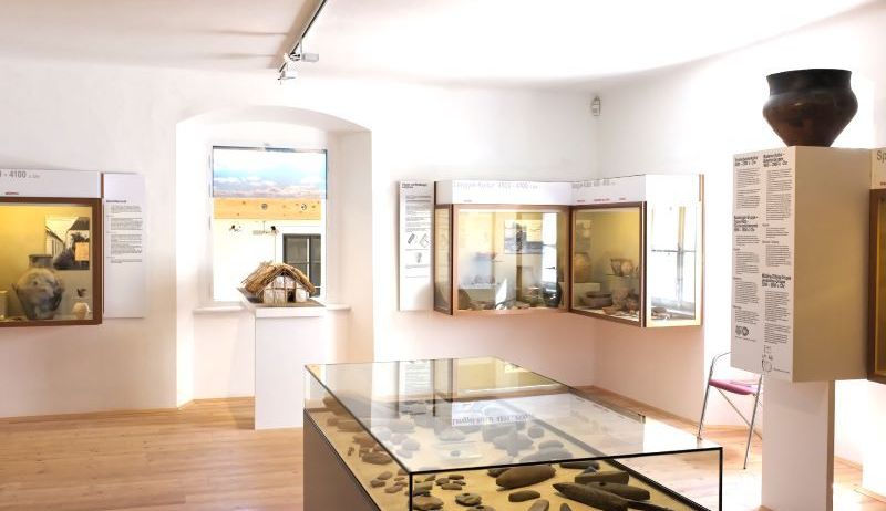 Ein Ausstellungsraum mit weißen Wänden und Holzböden. Verschiedene Artefakte sind in Glasvitrinen ausgestellt, darunter Keramik und andere Gegenstände. Ein Fenster an der Wand zeigt eine Landschaft.