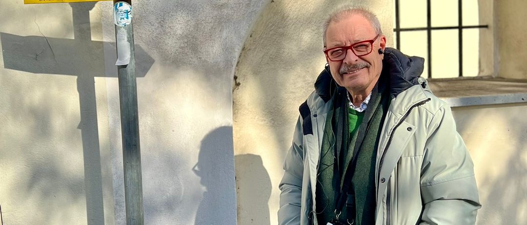 Ein Mann in einem grauen Mantel steht neben einem gelben Schild mit Wegbeschreibungen zu Rosenberg Rundweg und Maria Dreieichen Rundweg. Das Gebäude hat ein Fenster und einen Bogen.