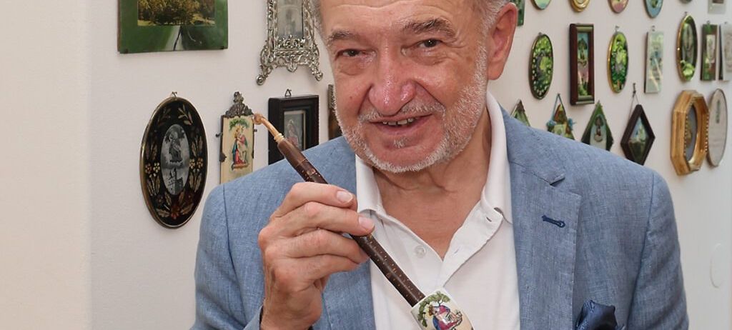 Ein älterer Mann steht vor einer Wand mit vielen gerahmten Bildern. Er lächelt und hält eine Pfeife in der Hand.