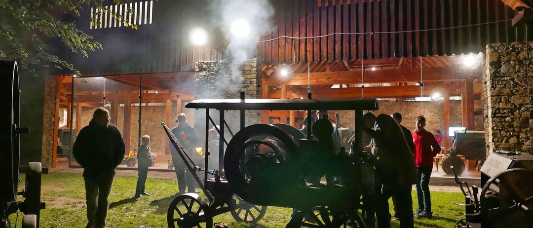 Eine Gruppe von Menschen versammelt sich um ein dampfbetriebenes Fahrzeug auf einem Rasenfeld bei Nacht, mit einem Holzgebäude im Hintergrund.