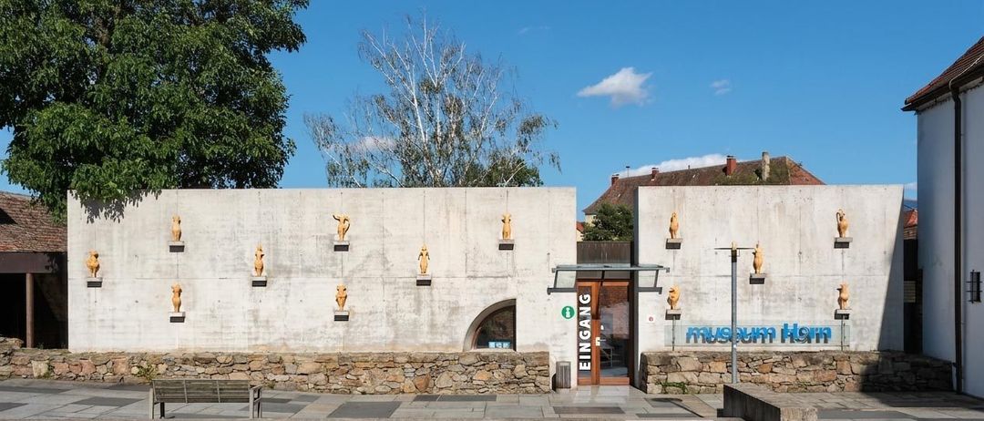 Eine Betonwand mit einer Reihe goldener Statuen und einem Schild mit der Aufschrift 'Eingang' über einer Holztür. Die Wand wird von Steinmauern flankiert, und ein Baum ist im Hintergrund sichtbar.