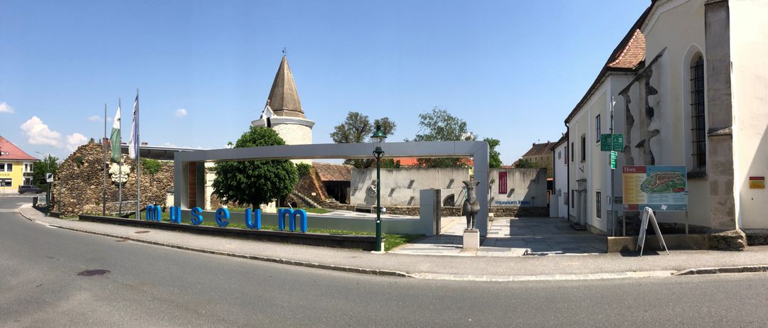 Der Eingang zu einem Museum ist durch ein großes blaues Schild mit der Aufschrift 'Museum' gekennzeichnet. Eine Statue steht vor dem Eingang, und eine Straßenlaterne ist in der Nähe. Hinter dem Museum befindet sich ein Gebäude mit einem Turm und Bäumen.