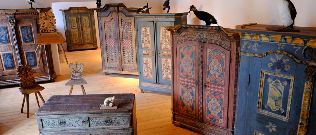 Ein Raum mit mehreren antiken Holzschränken und Schubladen, jeweils mit einzigartigen Designs. Ein Holzhocker und ein Holzschreibtisch stehen auf dem Boden. Ein Vogel sitzt auf einem der Schränke.