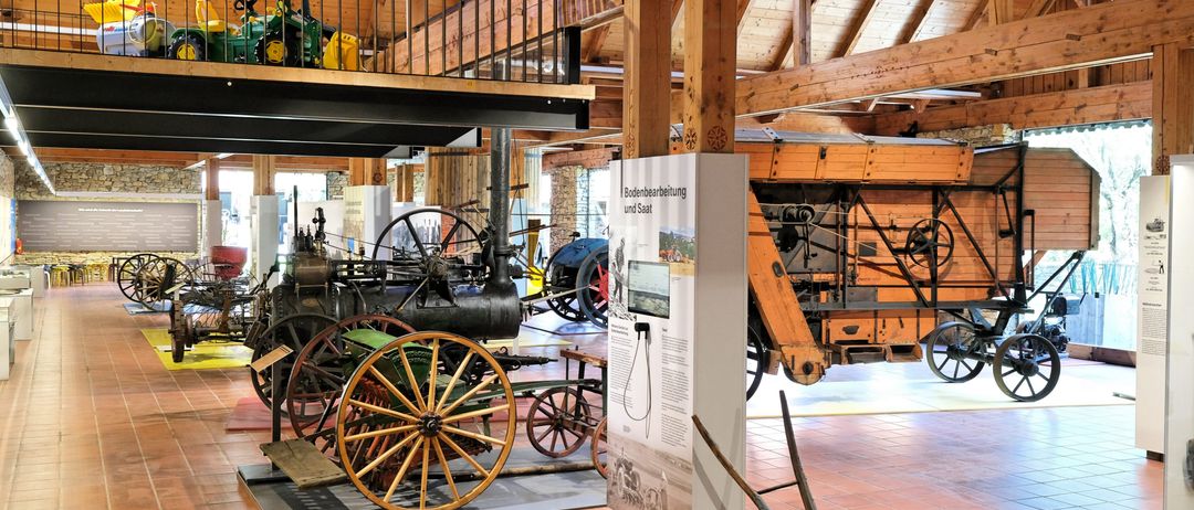 Eine Indoor-Museumsausstellung mit einer Sammlung von Oldtimern, darunter ein Traktor, eine Dampflokomotive und Pferdekutschen, ausgestellt auf einem gefliesten Boden unter Holzbalken.