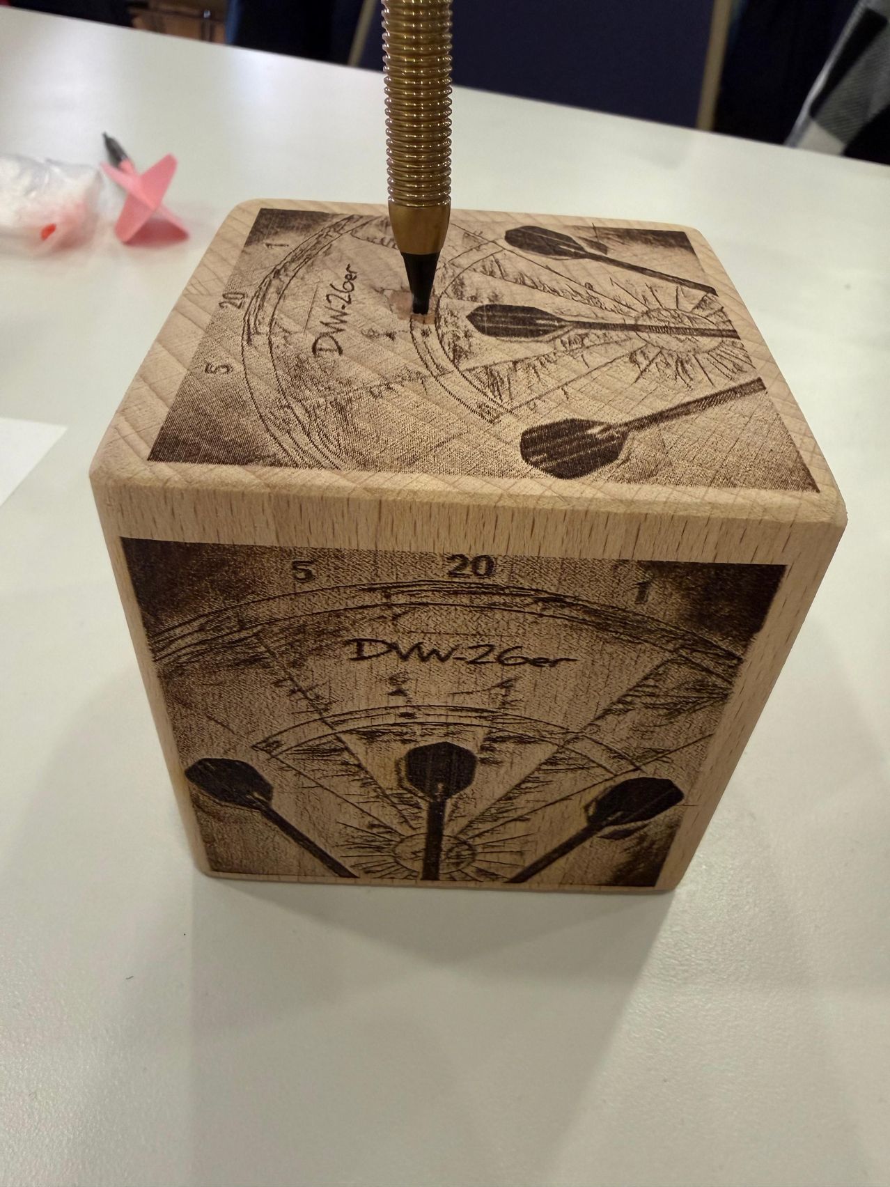 Ein Holzkasten mit einer Dartboard-Gravur. Darin steckt ein Dart. Der Kasten hat Nummern von 1 bis 20.