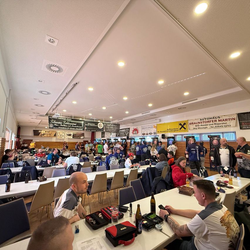 Ein großer Raum voller Menschen, die an Tischen sitzen und scheinbar ein Brettspiel spielen. Banner und Schilder hängen von der Decke. Zwei Männer sitzen eng beieinander und konzentrieren sich auf ein Brettspiel vor ihnen.