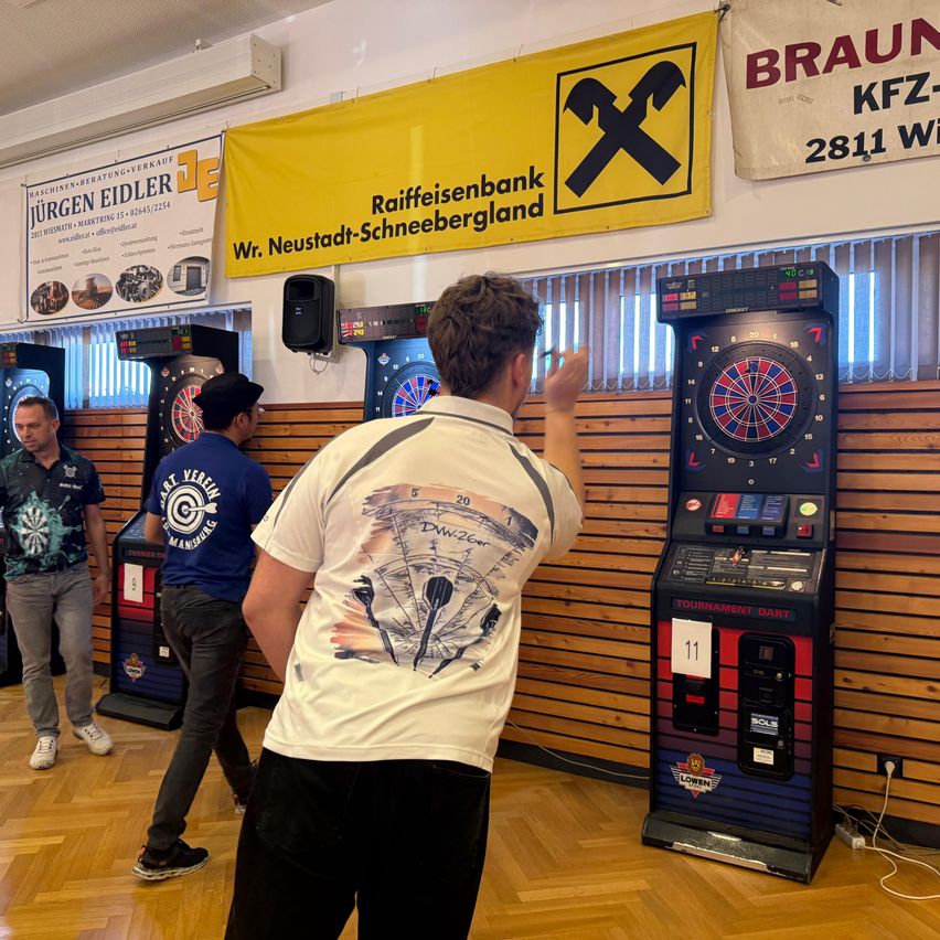 In einem Raum spielt ein Mann Darts, während zwei andere Männer zuschauen. Ein Dartboard ist an einer Holzwand montiert.