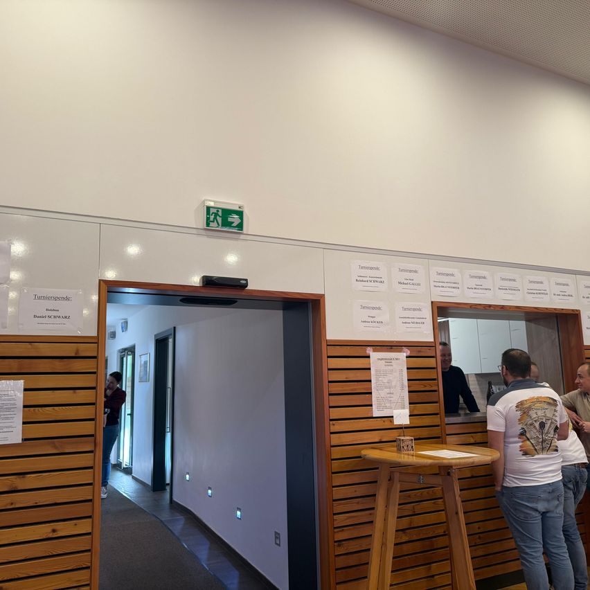 Ein Innenbereich mit Holzpaneelen und einer weißen Wand. Papiere sind an der Wand angebracht, und ein grünes Ausfahrtsschild befindet sich darüber. Vor einem Tisch stehen Personen, von denen eine eine Uhr trägt.