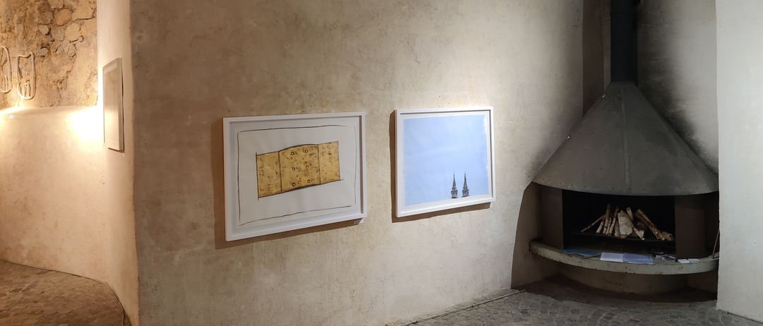 Ein kleiner Raum mit zwei gerahmten Bildern an der Wand, eines zeigt ein altes Buch und das andere eine Stadtlandschaft. Rechts befindet sich ein Kamin mit Holzscheiten, bereit zum Entzünden.