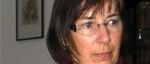 Eine ältere Frau mit kurzen Haaren und Brille trägt ein graues Oberteil und einen Weinroten Schal mit einem roten Anhänger. Sie steht in einem Raum.