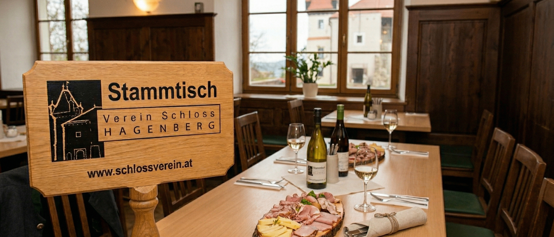 Ein Holzschild mit der Aufschrift Stammtisch. Ein Tisch ist mit Fleisch, Käse und Weingläsern gedeckt. Ein Fenster zeigt ein Gebäude mit Turm.