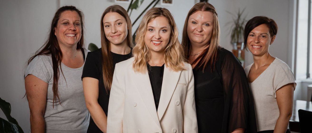 Fünf Frauen stehen zusammen und lächeln für ein Gruppenfoto. Die Frau in der Mitte trägt eine weiße Jacke und ein schwarzes Oberteil. Hinter ihr tragen drei Frauen schwarze Oberteile. Im Hintergrund sind eine Pflanze und Bilderrahmen zu sehen.