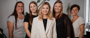 Five women stand together, smiling for a group photo. The woman in the center wears a white blazer and black shirt. Behind her, three women wear black tops. In the background, a plant and picture frames are visible.