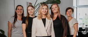 Fünf Frauen stehen lächelnd in einem Raum. Eine Frau in der Mitte trägt einen weißen Blazer. Hinter ihnen steht eine Pflanze, ein Glastisch und ein Fenster.