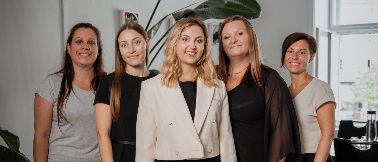 Fünf Frauen stehen lächelnd in einem Raum. Eine Frau in der Mitte trägt einen weißen Blazer. Hinter ihnen steht eine Pflanze, ein Glastisch und ein Fenster.