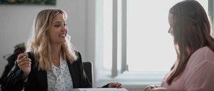 Zwei Frauen, eine lächelnd, sitzen an einem Tisch mit Papieren. Eine hält ein offenes Buch und einen Stift. Sie scheinen sich in einem Meeting zu befinden oder etwas zu besprechen.