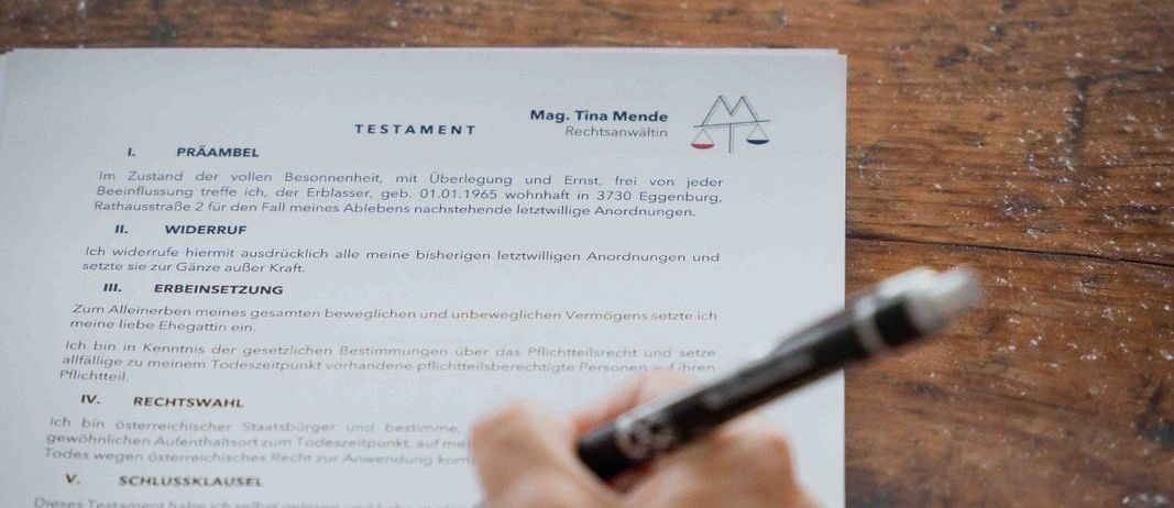 Eine Person füllt ein rechtliches Dokument mit dem Titel 'Testament' auf einem Holzschreibtisch aus. Das Dokument ist in deutscher Sprache, mit Abschnitten wie 'Praambel' und 'Widerruf'. Ein Stift ist in der Hand der Person.