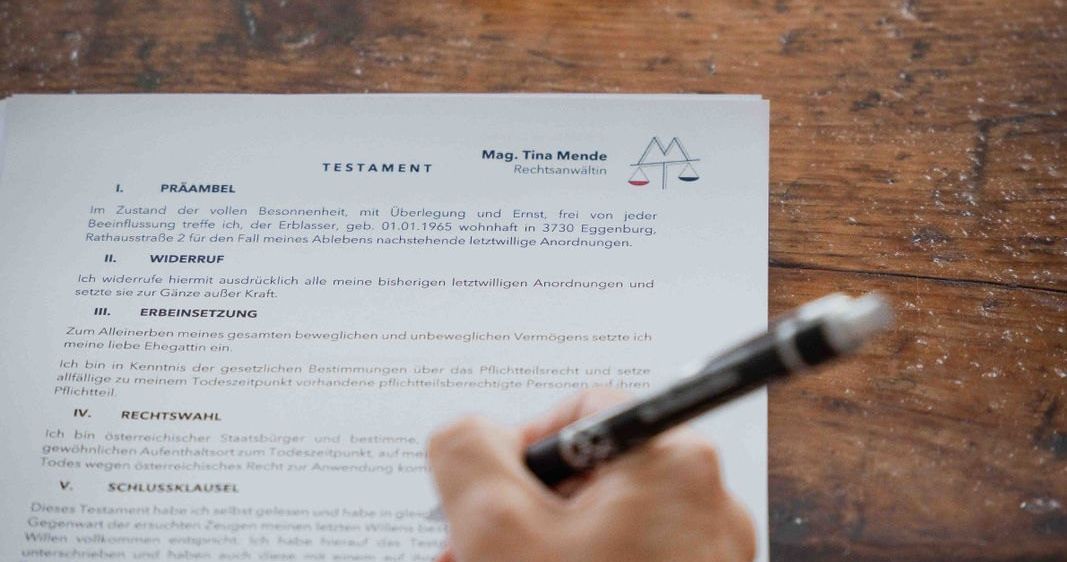 Eine Person füllt ein rechtliches Dokument mit dem Titel 'Testament' auf einem Holzschreibtisch aus. Das Dokument ist in deutscher Sprache, mit Abschnitten wie 'Praambel' und 'Widerruf'. Ein Stift ist in der Hand der Person.