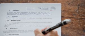 Eine Person füllt ein rechtliches Dokument mit dem Titel 'Testament' auf einem Holzschreibtisch aus. Das Dokument ist in deutscher Sprache, mit Abschnitten wie 'Praambel' und 'Widerruf'. Ein Stift ist in der Hand der Person.