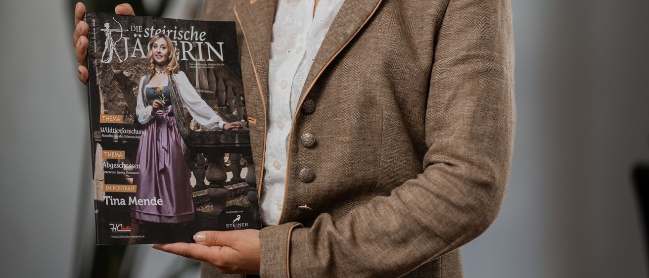 Eine Frau hält eine Zeitschrift mit einer Frau in traditioneller Kleidung, die lächelt, vor grauem Hintergrund.