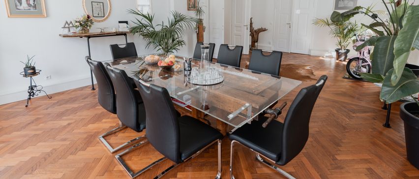 Ein moderner Essbereich mit einem Glastisch, schwarzen Stühlen und Holzboden. Die Einrichtung umfasst Pflanzen, Spiegel und gerahmte Bilder.