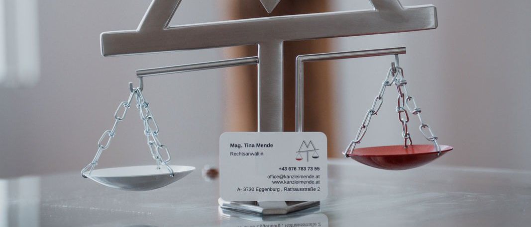 Eine silberne und rote Waage mit einem Visitenkarte, das Mag. Tina Mende liest, ist auf einer Glasoberfläche platziert. Die Waage hat eine dreieckige Form mit einer roten Kugel unten. Die Visitenkarte hat eine Telefonnummer und eine Website-Adresse.