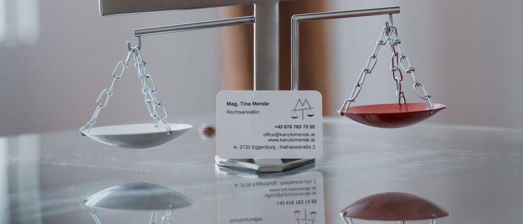 A silver and red scale with a business card labeled Mag. Tina Mende is placed on a glass surface. The scale has a triangular shape with a red ball at the bottom. The business card has a contact number and website address.