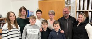 Eine Gruppe von sieben Personen, darunter fünf Kinder und zwei Erwachsene, posiert für ein Foto in einem Raum. Hinter ihnen befindet sich ein Regal mit Büchern und eine Tafel.