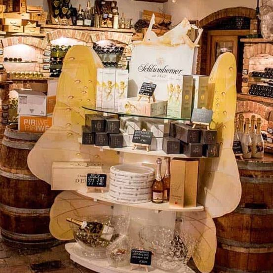 Ein Weinladen zeigt Flaschen, Kisten und Teller auf einem großen Regal vor hölzernen Fässern. Das Regal hat ein gelbes Design. Oben befinden sich Weinflaschen und Kisten an einer Ziegelwand.