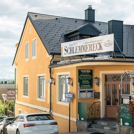 Ein Gebäude mit schwarzem Dach und einem Schild, das 'Zwetller Emmereck' steht. Das Gebäude hat eine gelbe Fassade mit Fenstern und einem Holzzaun mit roten Blumen.