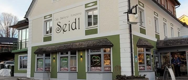 Die Fassade eines Gebäudes namens Seidl, mit einem Café-Schild, Fenstern und einem Schornstein, unter einem klaren blauen Himmel.