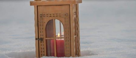 Eine hölzerne Laterne mit einer roten Kerze darin, teilweise im Schnee vergraben. Die Laterne hat komplizierte Muster an ihren Seiten.