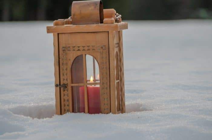 Eine hölzerne Laterne mit einer roten Kerze darin, teilweise im Schnee vergraben. Die Laterne hat komplizierte Muster an ihren Seiten.