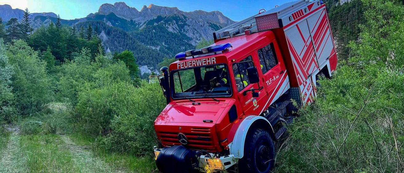 Ein roter Feuerwehrwagen steckt im Gras fest, mit einem Gebirgszug im Hintergrund.